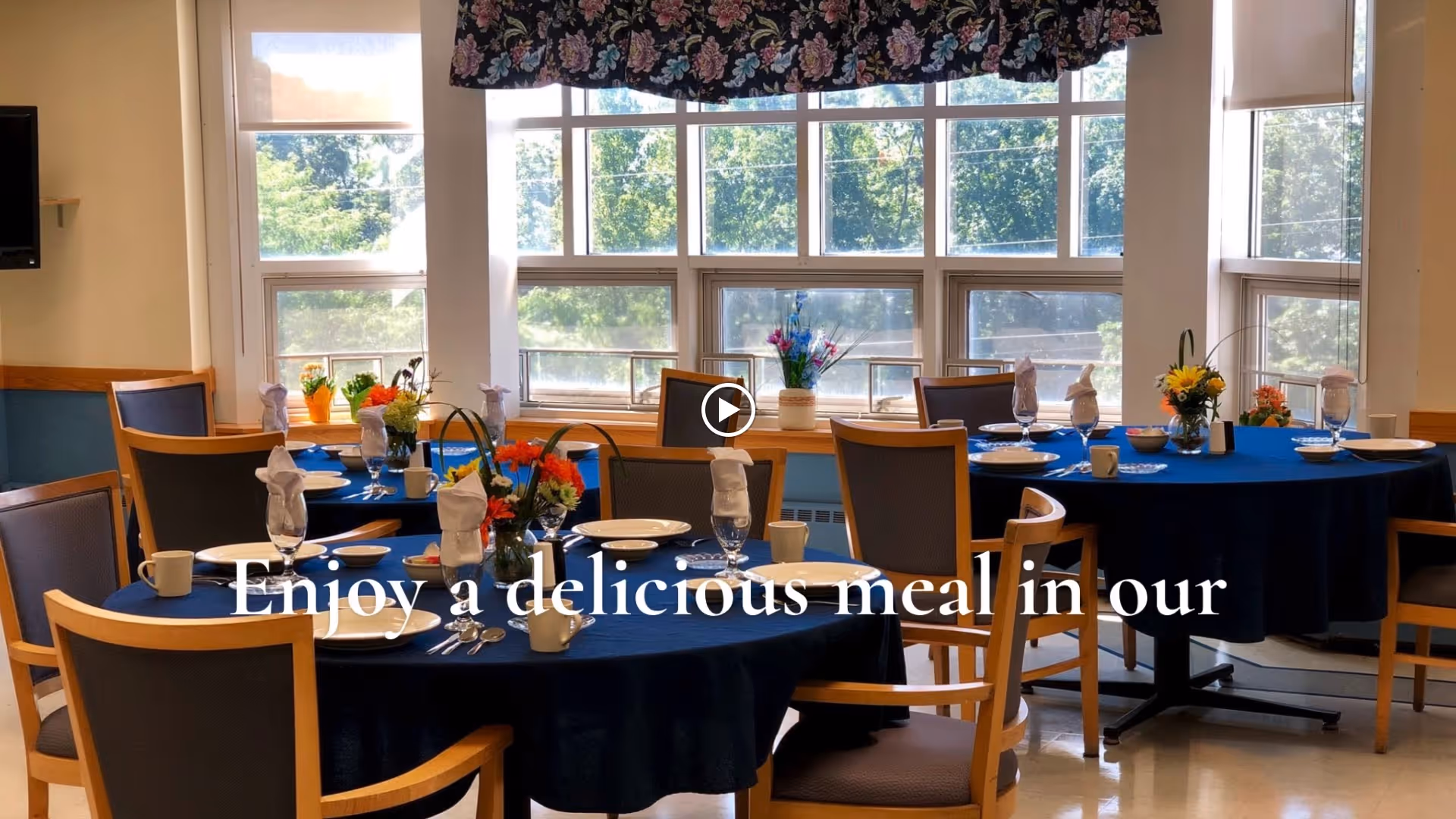 Dining room with round tables covered in dark blue tablecloths, set with plates, glasses, napkins, and silverware. Each table has a floral centerpiece. Large windows in the background let in natural light and show green trees outside.