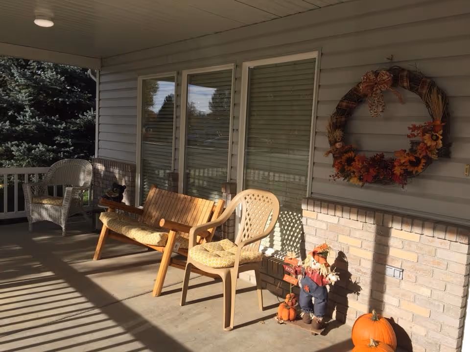A covered porch area with a wooden bench and two chairs, one wicker and one plastic, all with cushions. The porch has a brick half-wall and siding with three large windows. There is a fall-themed wreath hanging on the wall and autumn decorations including a scarecrow and pumpkins on the floor. Sunlight casts shadows from the porch railing onto the concrete floor.