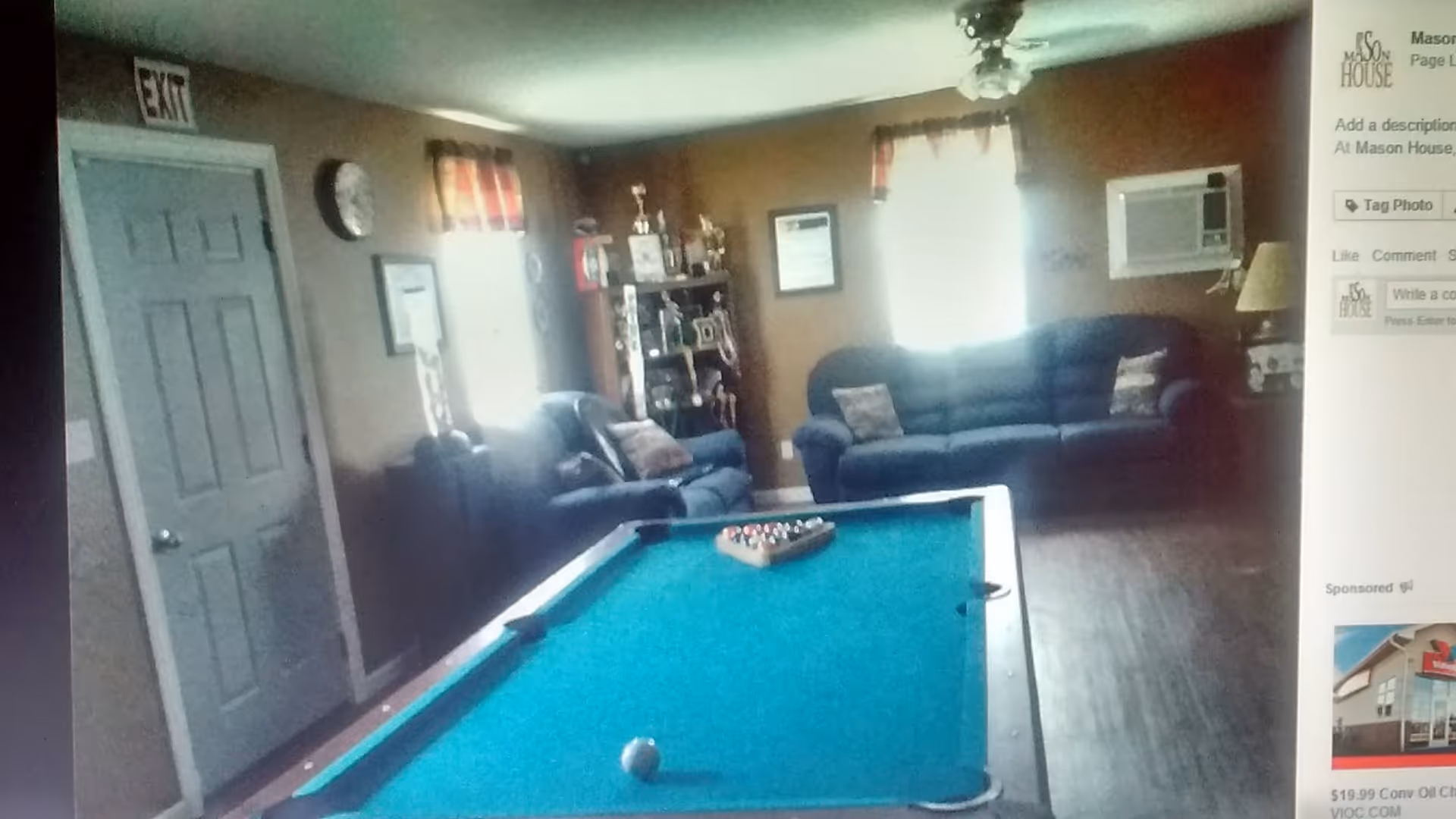 Interior room with a pool table in the foreground, two dark blue sofas against the walls, a clock and framed pictures on the walls, a ceiling fan, and a window with curtains letting in light.