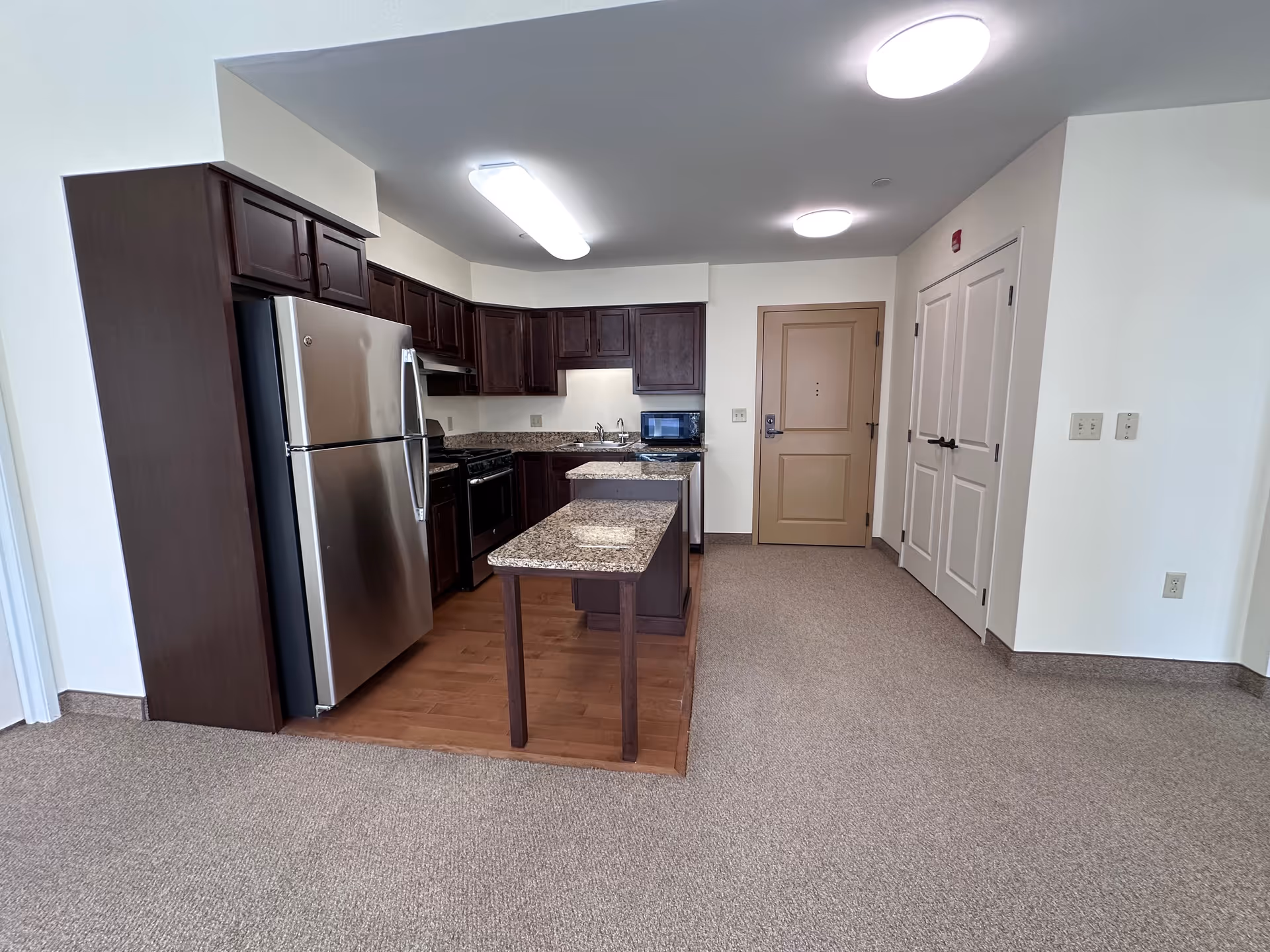 Interior view of a kitchen area in a senior living facility with dark wood cabinets, a stainless steel refrigerator, a stove, a microwave, and granite countertops. The kitchen has a small island with an extended countertop supported by a leg. The floor in the kitchen area is wood, while the surrounding area is carpeted. There is a beige door and a double-door closet on the right side of the image.