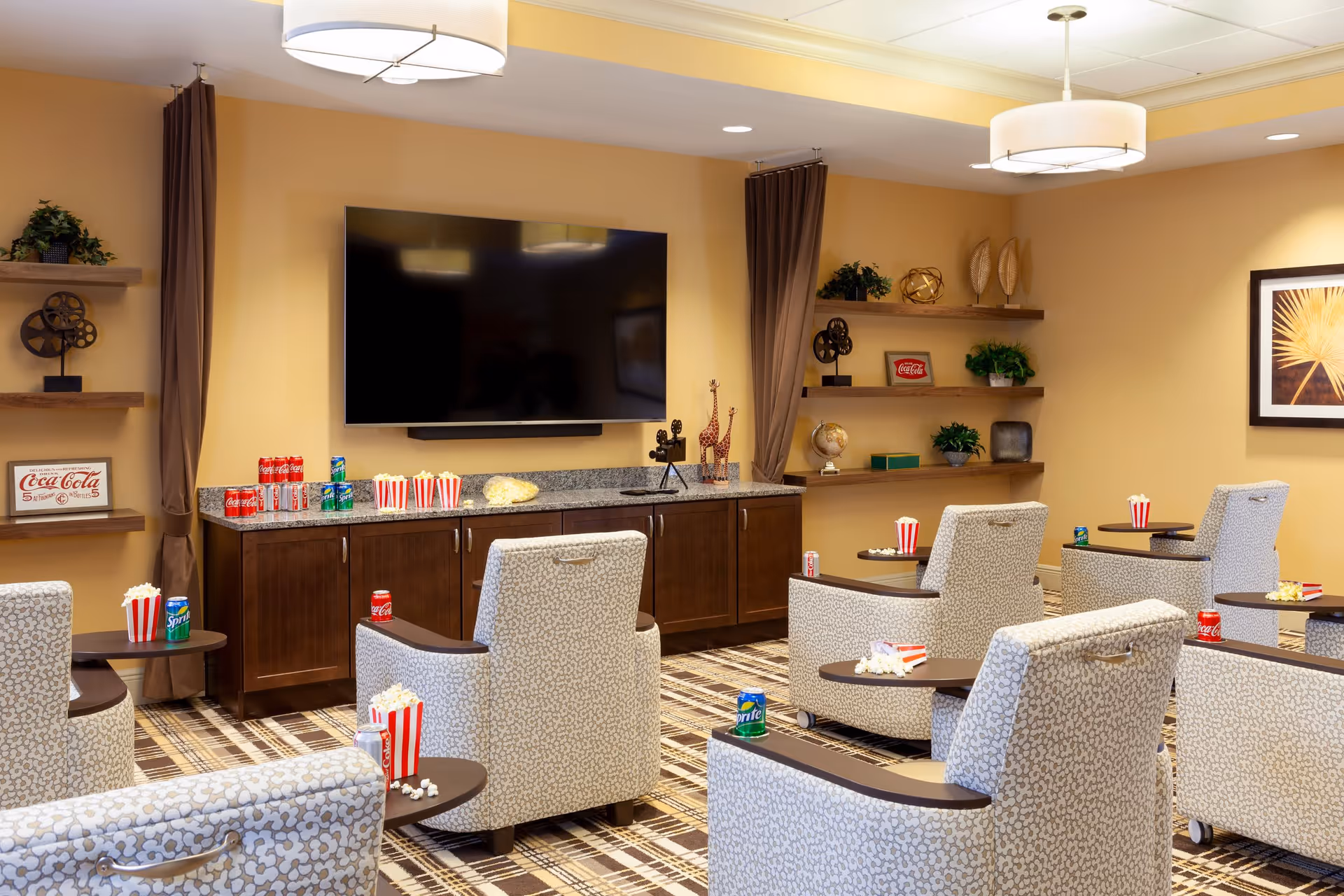 A cozy entertainment room with patterned armchairs arranged in rows facing a large flat-screen TV mounted on a beige wall. Each chair has a small side table holding popcorn and soda cans. The room features wooden shelves with decorative items, including plants, giraffe figurines, and vintage Coca-Cola signs. The floor is carpeted with a plaid pattern, and two ceiling lights provide warm illumination.