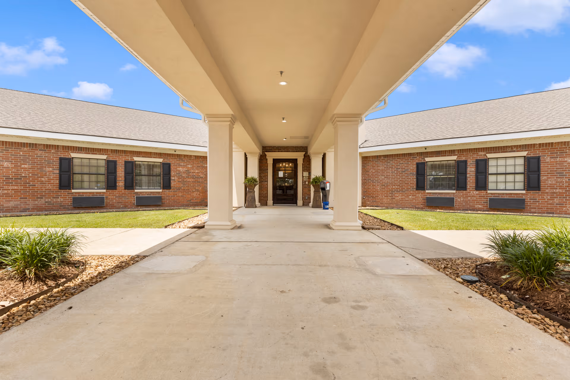 Covered walkway entrance to a brick building with black window shutters, green grass, and landscaped plants on either side under a blue sky with some clouds.