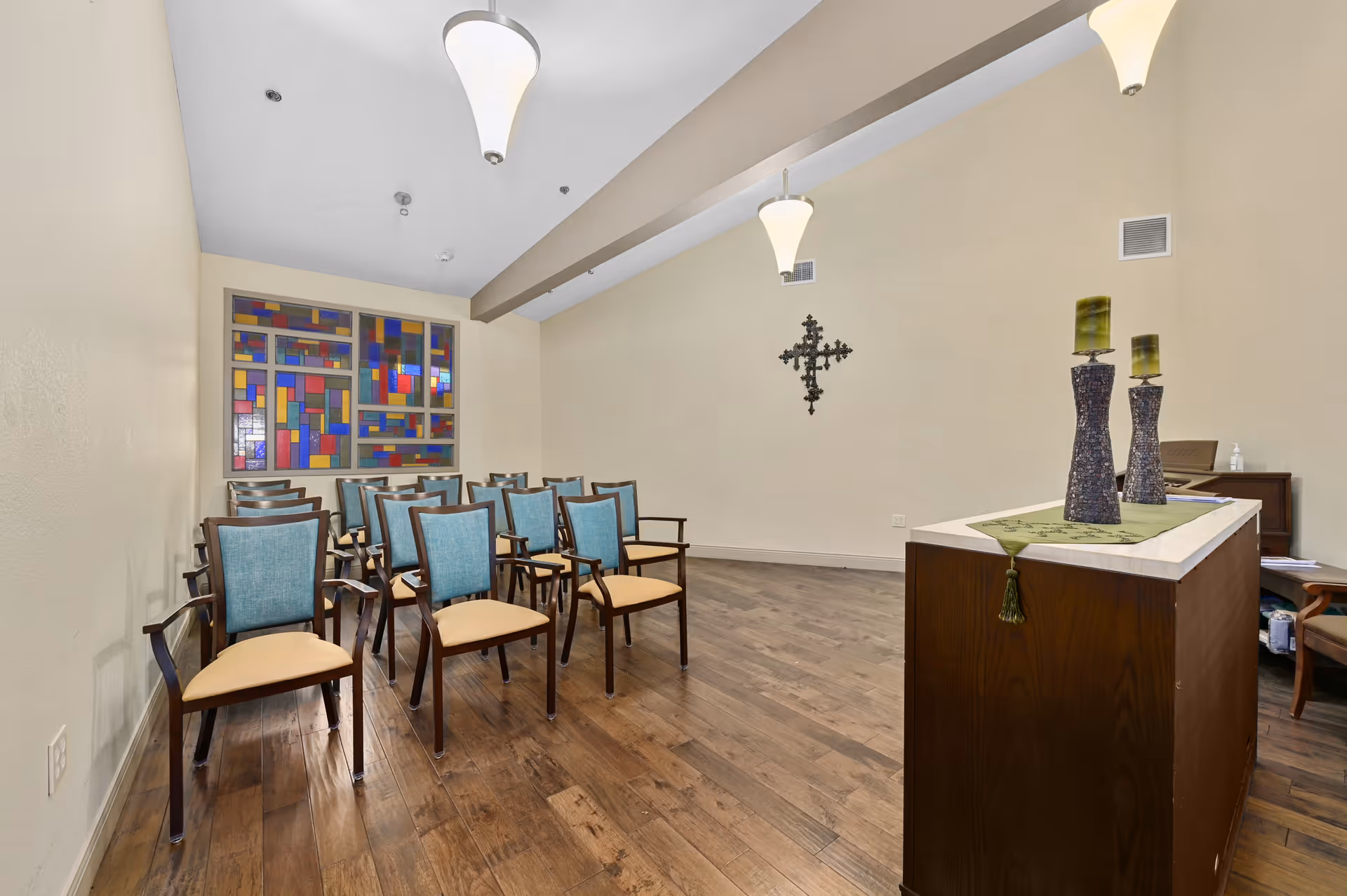 A small chapel or meditation room with wooden floor and beige walls. There are two rows of wooden chairs with blue backs and beige seats facing a wooden podium adorned with two tall decorative candle holders and a green runner. On the back wall, there is a colorful stained glass window and a decorative metal cross mounted on the right wall. Three pendant lights hang from the ceiling.