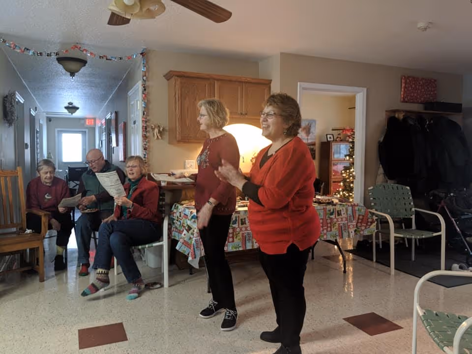A group of elderly people gathered in a common area of an assisted living facility. Two women stand in the foreground, one clapping and smiling, while three others sit on chairs along the hallway wall, looking at papers and engaging with each other. The room is decorated with holiday-themed tablecloths and a Christmas tree is visible in an adjacent room. Coats hang on a rack near the entrance.
