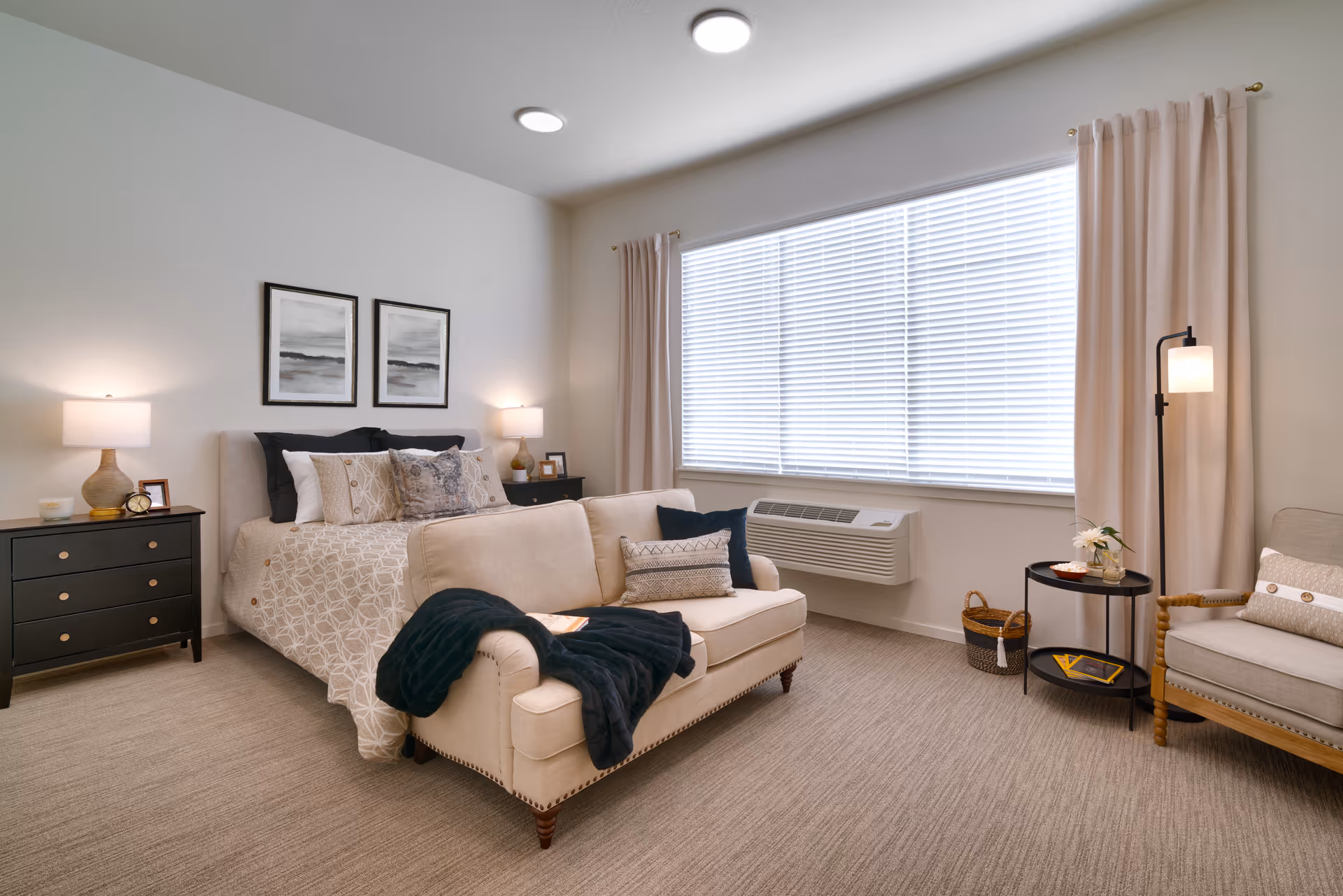 A well-lit bedroom featuring a bed with patterned bedding and multiple pillows, flanked by two black nightstands each with a lamp and decorative items. At the foot of the bed is a beige upholstered loveseat with a dark throw blanket and pillows. A large window with closed blinds and beige curtains is on the right wall, beneath which is a wall-mounted air conditioning unit. To the right of the loveseat is a round black side table with a lamp, a small plant, and a basket on the floor next to it. A beige armchair with a pillow is positioned near the side table. The room has beige carpeting and neutral-colored walls.