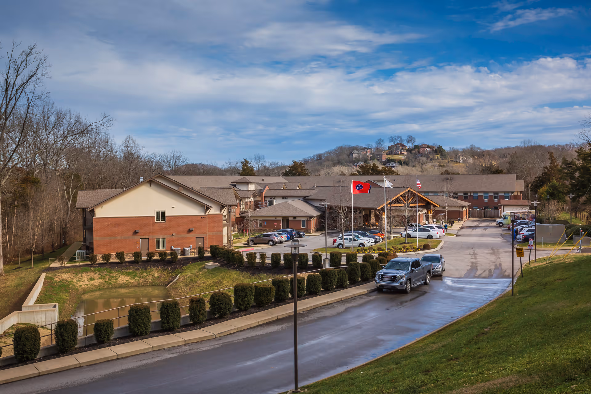 Exterior view of The Lodge at Natchez Trace, showing a large brick and beige building with a parking lot in front. Several cars are parked and driving on the road leading to the entrance. There are three flagpoles with flags flying near the entrance. The surrounding area includes trees, grassy hills, and a partly cloudy sky.