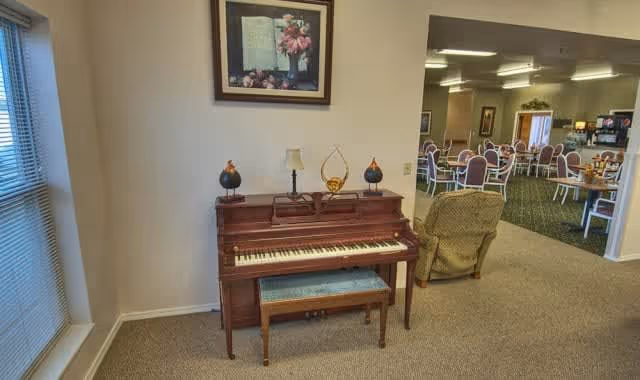 Interior view of a senior living facility showing a wooden piano with a bench in front of it, decorated with two small lamps and a decorative item on top. Above the piano hangs a framed floral artwork. To the right, there is a green upholstered armchair and an open area leading to a dining room with multiple round tables and chairs. The room is well-lit with ceiling lights and has carpeted floors.