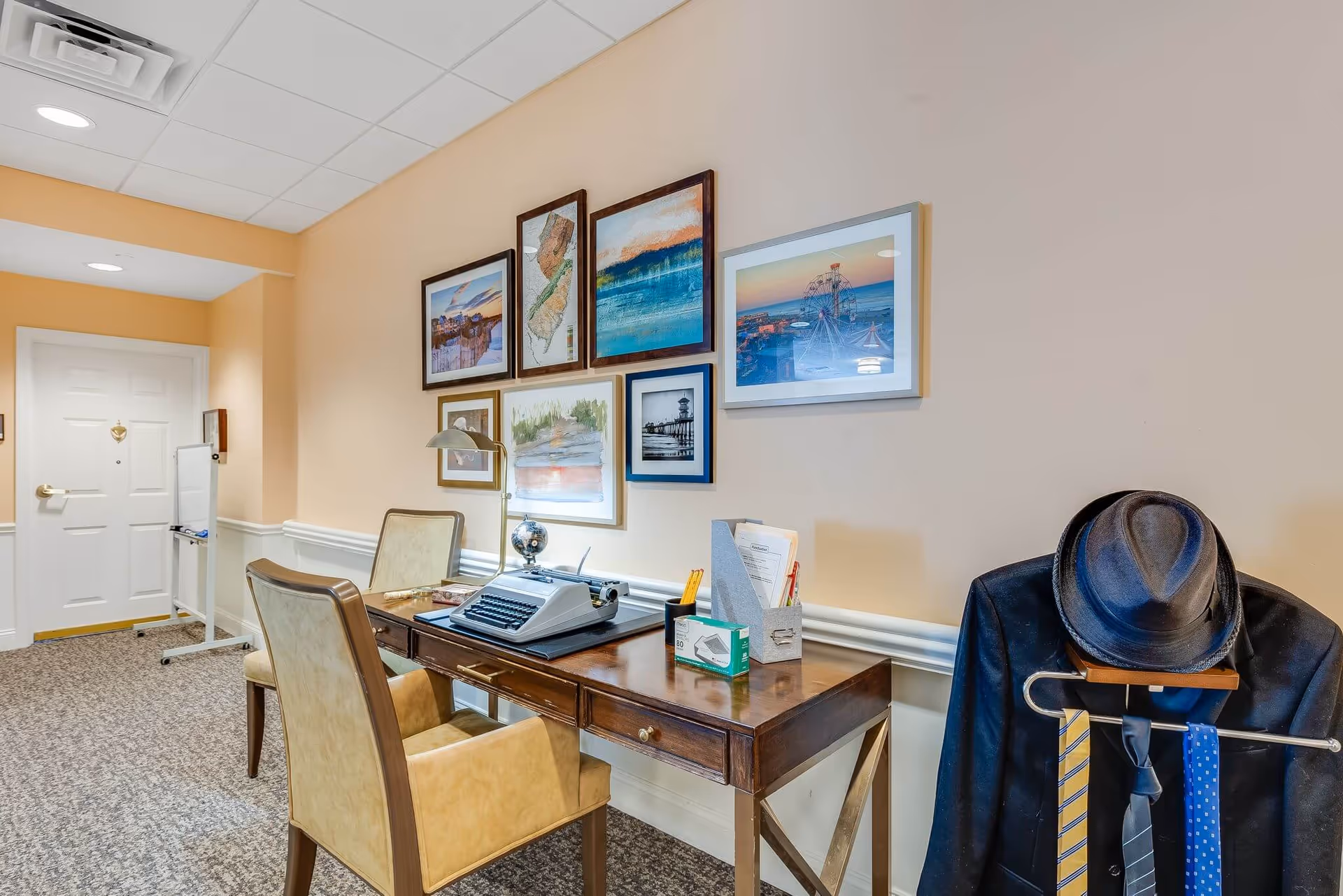 A cozy interior corner of a senior living facility featuring a wooden desk with a vintage typewriter, a desk lamp, and office supplies. Two beige upholstered chairs are positioned at the desk. The wall above the desk is decorated with framed pictures and artwork. To the right, a coat rack holds a dark jacket, a hat, and several ties. The floor is carpeted, and a white door is visible in the background.