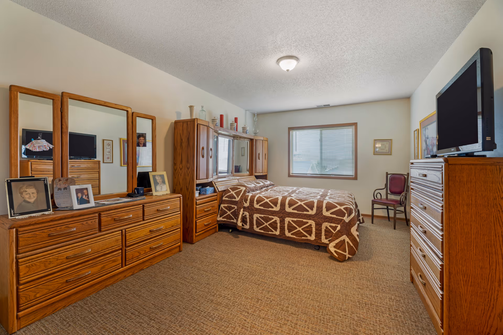 A senior living bedroom with a bed covered in a brown and beige patterned quilt. The room features wooden furniture including a dresser with three mirrors, a tall chest of drawers with a TV on top, and a headboard with cabinets. There are framed photos and decorative items on the dresser and walls. A window with blinds allows natural light into the room, and a single chair is placed near the window.