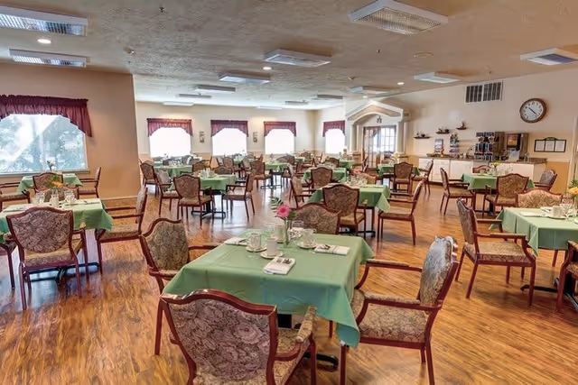 A spacious dining room with multiple tables covered in green tablecloths, each set with cups, plates, and napkins. The room has wooden flooring, floral upholstered chairs, large windows with maroon valances, and a clock on the wall. The room is well-lit with ceiling lights.