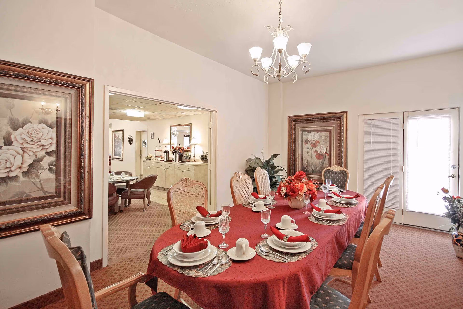 A dining room with a long table covered in a red tablecloth set for eight people with white plates, cups, silverware, and red napkins. There is a floral centerpiece on the table. The room has beige walls, framed floral artwork, a chandelier, and a door with blinds leading outside. Through an open doorway, another room with additional seating and a buffet area is visible.