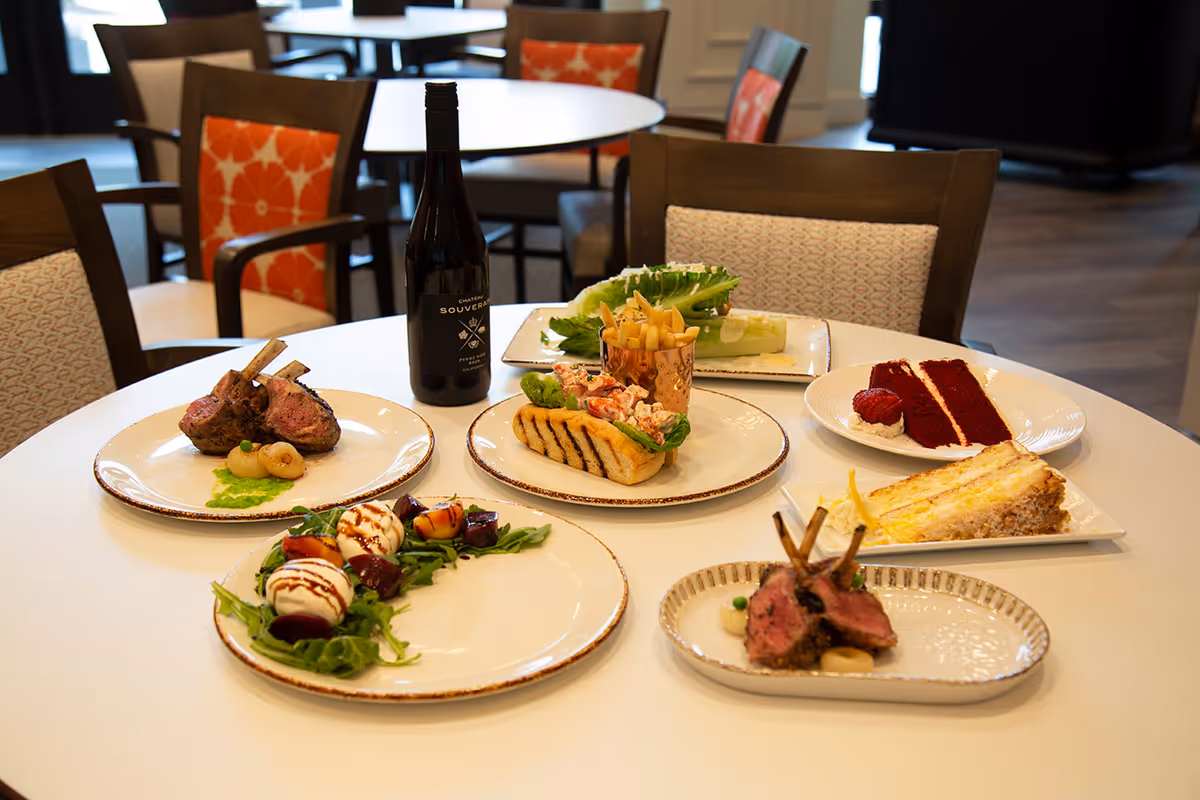 A round dining table set with various plated dishes including lamb chops, a lobster roll with fries, a salad with mozzarella and beets, two slices of cake, and a bottle of red wine. The background shows chairs with patterned cushions and a wooden floor.