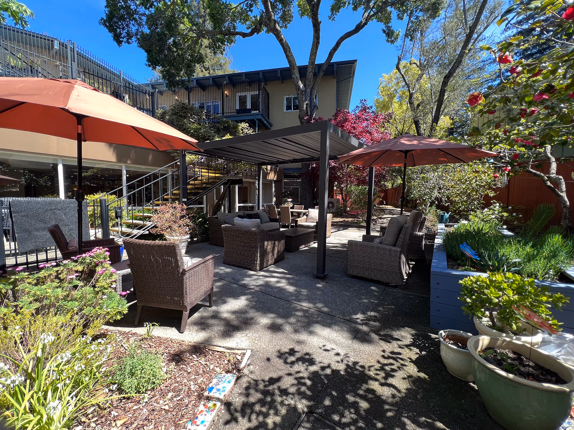 Outdoor patio area at WindChime of Marin Memory Care featuring wicker seating with cushions under two large red umbrellas and a pergola. The space is surrounded by various plants, flowers, and trees with a building and staircase in the background under a clear blue sky.