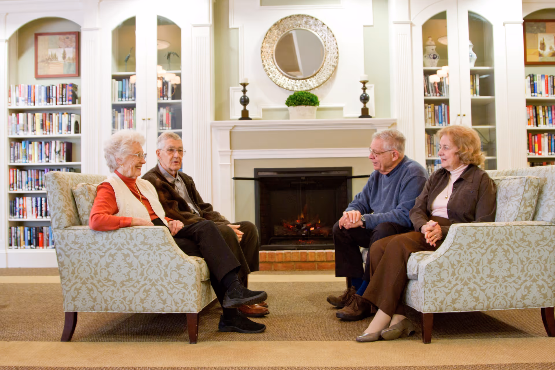 Four elderly people sitting on two patterned sofas facing each other in a cozy living room with a fireplace, a round decorative mirror above the mantle, and bookshelves filled with books on either side.