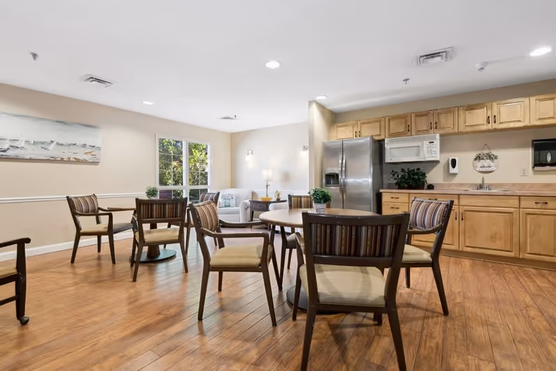 A bright and spacious common area with wooden flooring featuring several round tables surrounded by wooden chairs with striped cushions. In the background, there is a kitchen area with wooden cabinets, a stainless steel refrigerator, a microwave, and a sink. A window lets in natural light, and a white armchair with a side table and lamp is visible near the window. The walls are painted light beige, and a painting hangs on one wall.