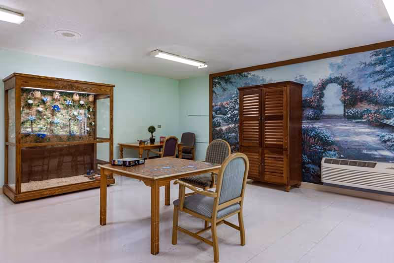 A room with a wooden table and four chairs arranged around it, a large wooden cabinet, and a decorative display case with flowers. The walls are light green, and one wall features a large mural of a garden pathway with an archway and flowers. There is also an air conditioning unit below the mural.