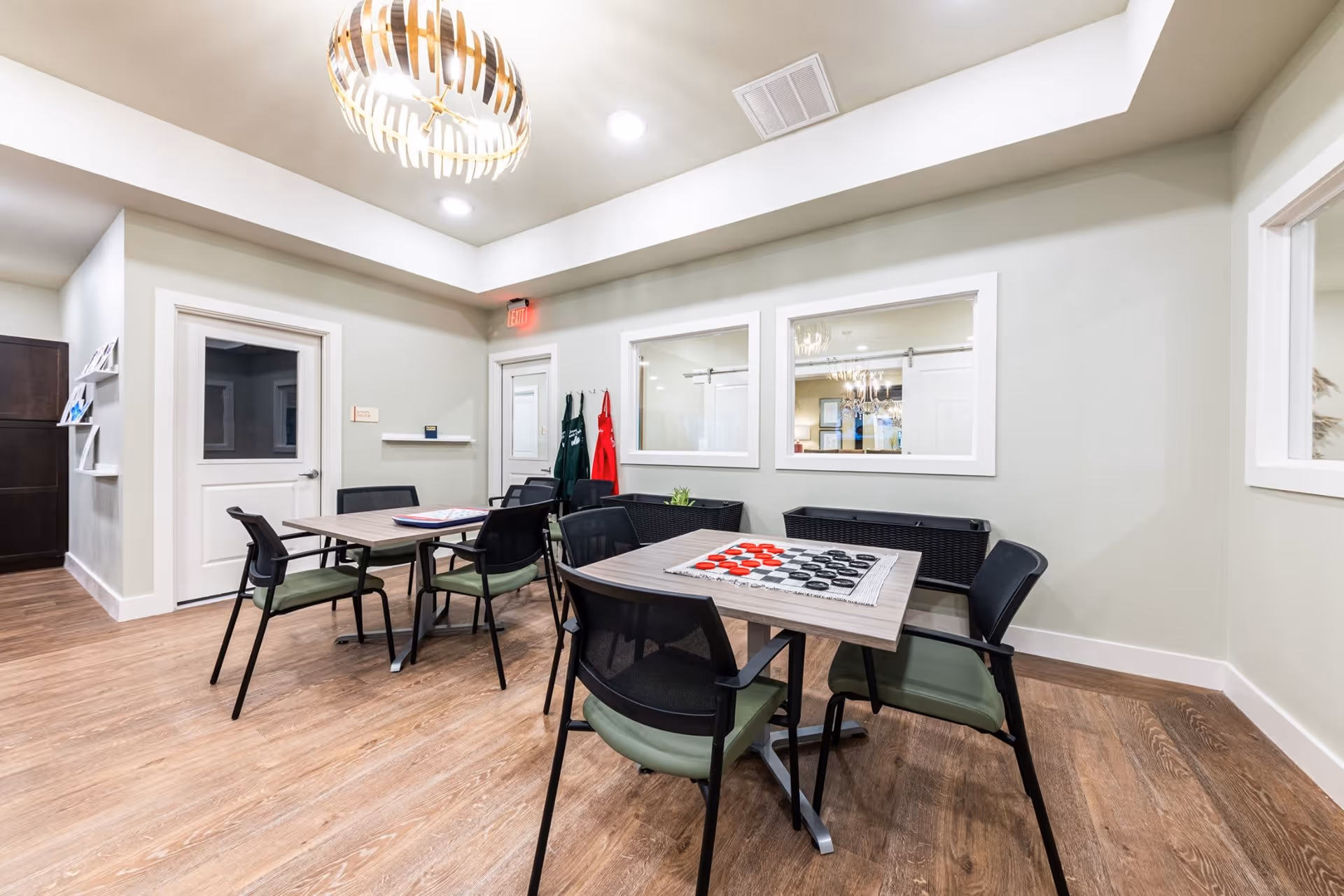 A well-lit room with wooden flooring featuring two tables with chairs around them. One table has a checkers game set up. The walls are light-colored with two large windows and a door. There are aprons hanging on hooks near the door and a modern light fixture on the ceiling.