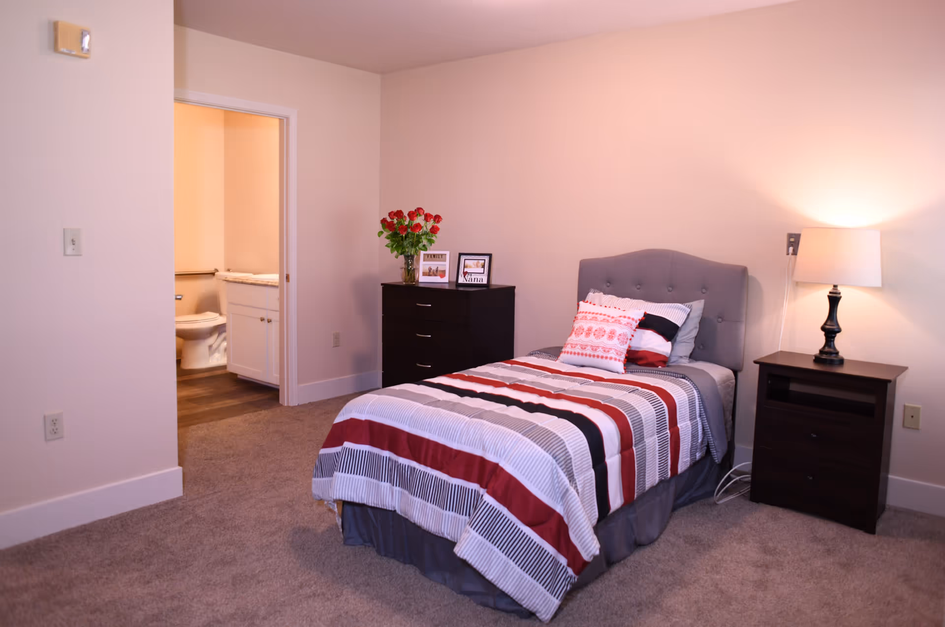 A bedroom with a single bed featuring a striped red, white, and gray comforter and decorative pillows. To the right of the bed is a dark wooden nightstand with a lamp. To the left of the bed is a dark wooden dresser with a vase of red roses and two framed photos. An open door reveals a bathroom with a toilet and a white vanity.
