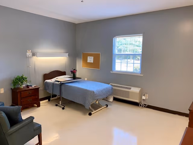 A simple senior bedroom with a single hospital-style bed, bedside table, armchair, window, and neutral gray walls.