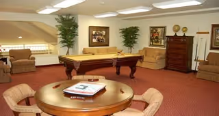 A cozy common area with a pool table in the center, surrounded by beige sofas and armchairs. There is a round wooden table with chairs in the foreground, and framed artwork and tall plants decorate the walls. The room has a warm red carpet and soft overhead lighting.