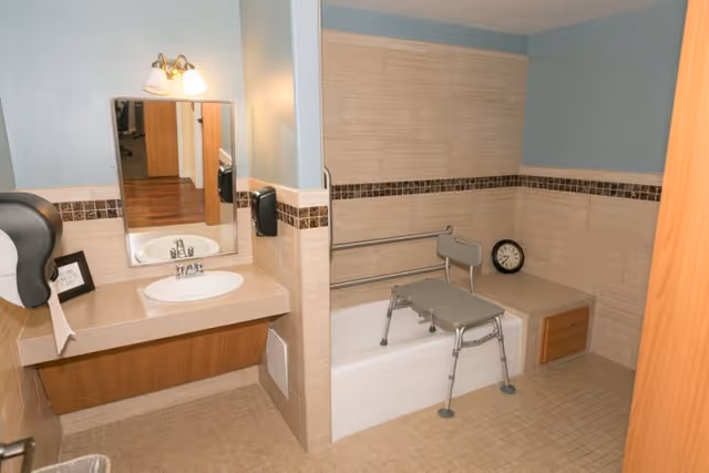A bathroom with beige tiled walls and floor, featuring a sink with a mirror above it, a mounted soap dispenser, a bathtub with safety grab bars, and a shower chair inside the tub. There is also a clock placed on a ledge next to the bathtub.