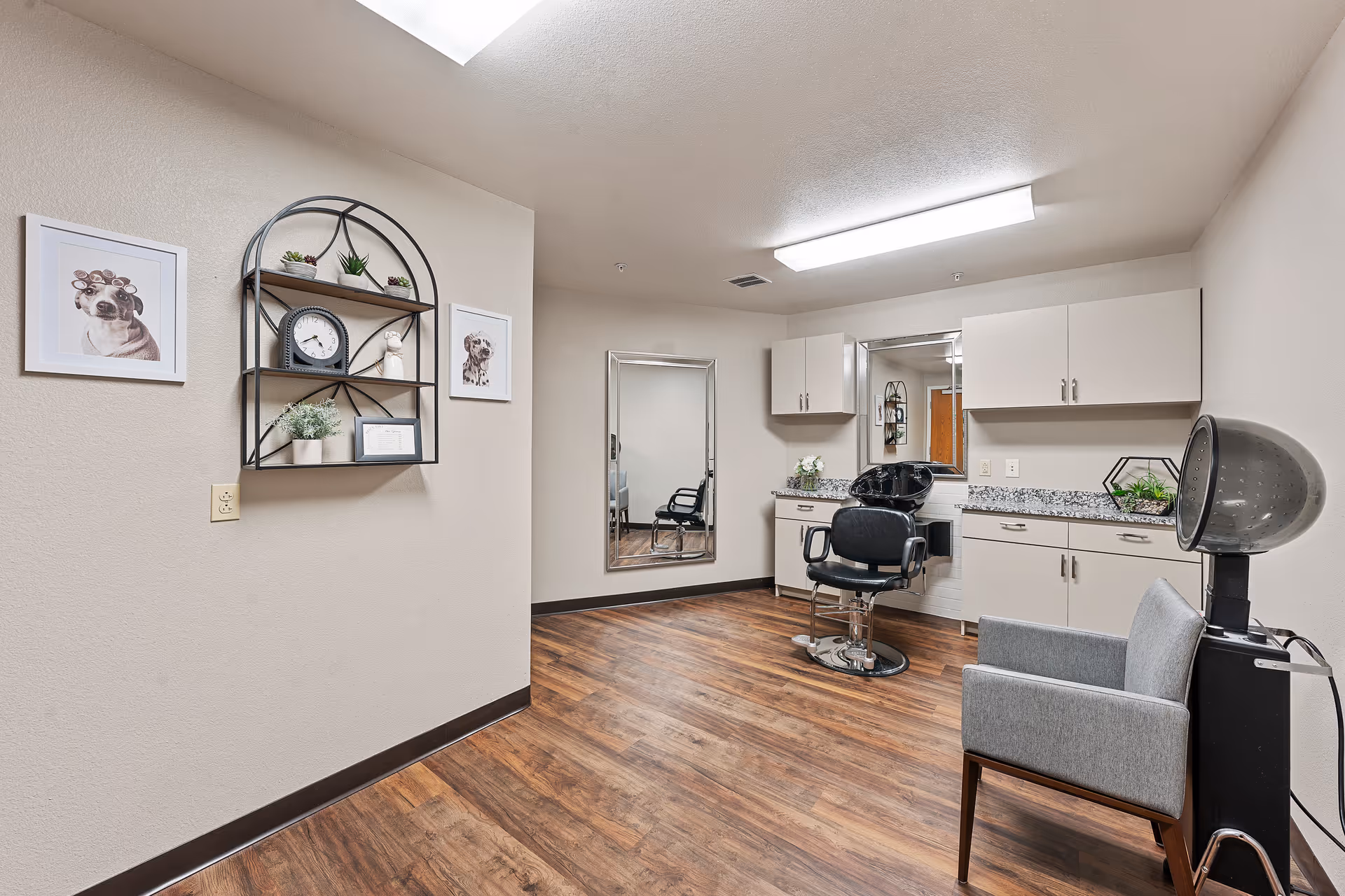 Interior view of a hair salon area in a senior living facility with a black salon chair, a hair washing station, a gray armchair, and a hair dryer. The room has wood flooring, beige walls, white cabinets with granite countertops, a large mirror, and decorative wall shelves with plants and framed pictures of dogs.