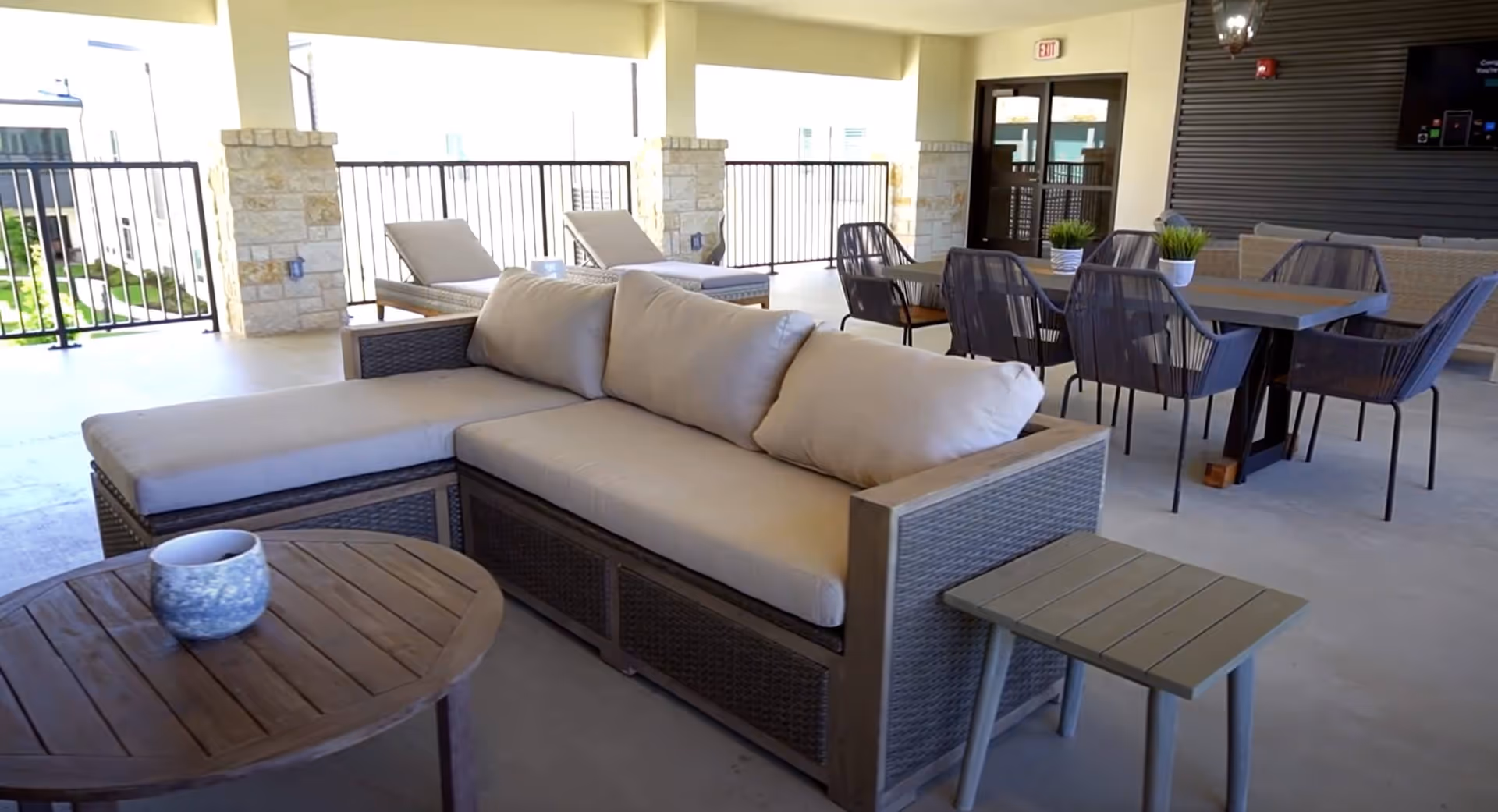 Covered outdoor patio area with cushioned wicker sectional sofa, round wooden coffee table with a small decorative pot, two lounge chairs, a dining table with six chairs, and potted plants on the tables. Stone pillars and metal railings surround the space.