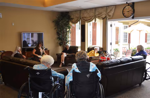 Elderly residents seated on sofas and in wheelchairs socializing in a communal living room with a TV and an open door to a patio.