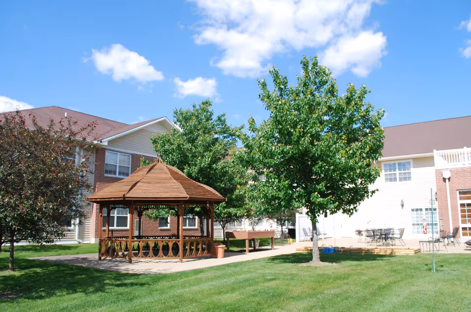 Outdoor area of Brookstone Estates of Robinson featuring a wooden gazebo, green trees, a grassy lawn, and a building with brick and siding in the background under a blue sky with some clouds.