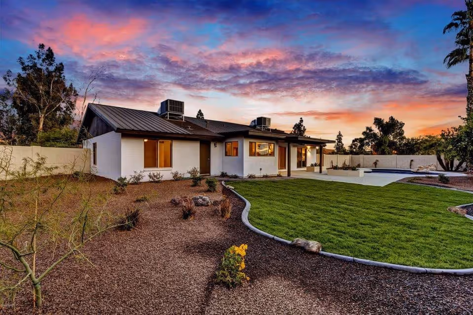 A single-story white building with a dark roof, surrounded by a landscaped yard featuring green grass, small plants, and trees under a colorful sunset sky.