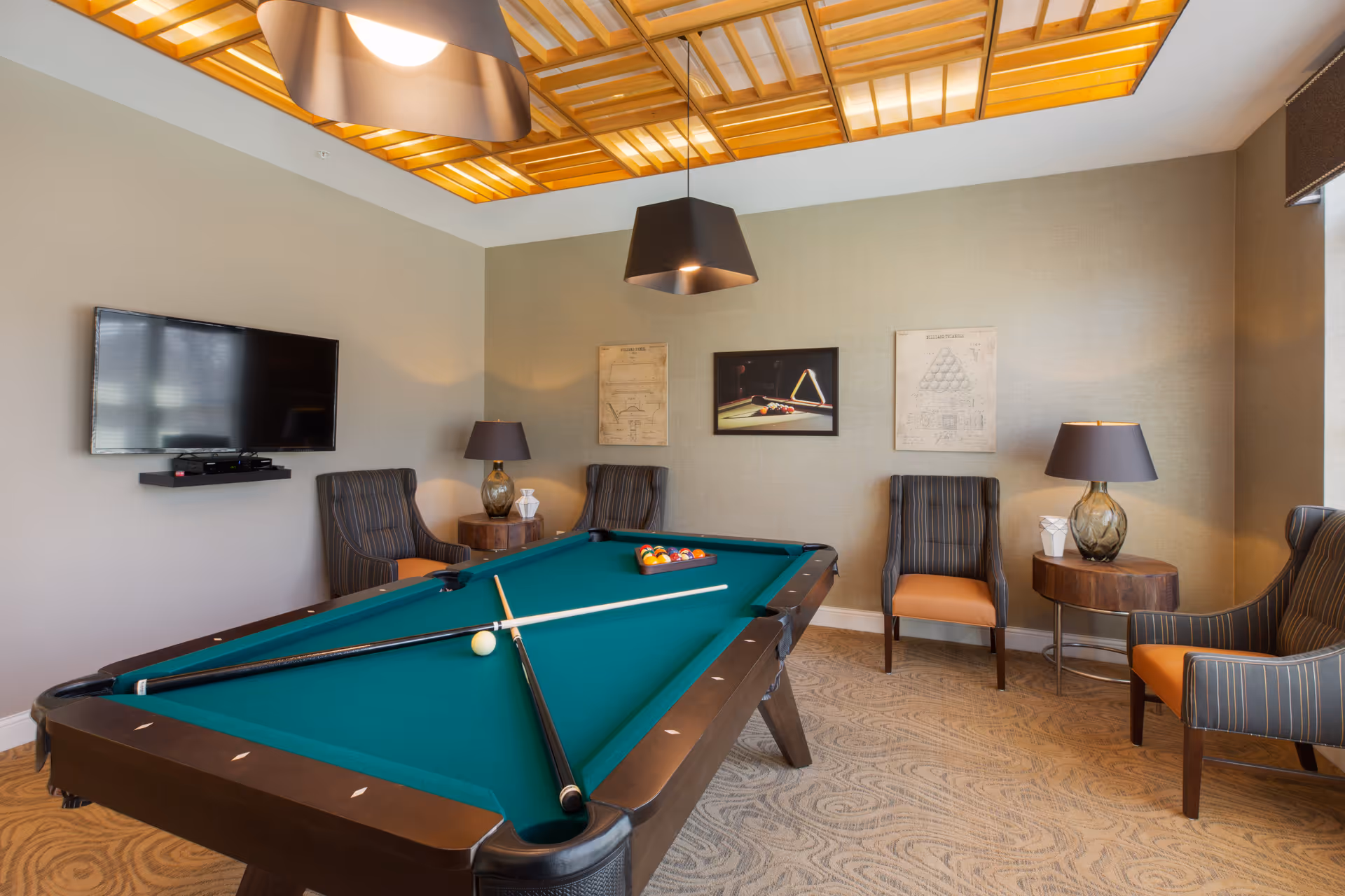 A well-lit game room featuring a pool table with two cues and a rack of billiard balls. The room has four striped armchairs with orange cushions, two side tables with lamps, a wall-mounted flat-screen TV, and framed billiards-themed artwork on the wall. The ceiling has a decorative wooden grid with warm lighting.
