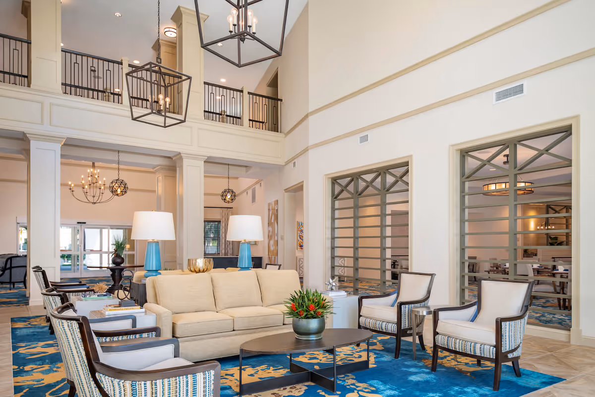 Spacious two-story common lounge with a beige sofa, armchairs, coffee table, blue patterned rug, and hanging chandeliers.