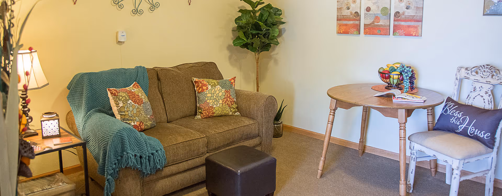 Cozy living room with a brown sofa draped with a teal throw, decorative pillows, a small ottoman, and a round wooden table with a chair and decor.