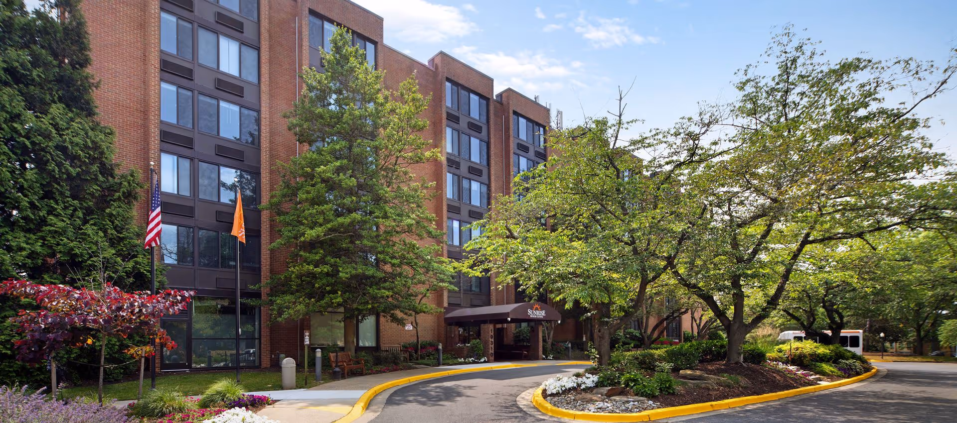 Exterior view of a multi-story brick building with large windows, surrounded by trees and landscaped greenery. There is a curved driveway with a yellow curb leading to an entrance canopy labeled 'Sunrise Senior Living'. Two flagpoles with an American flag and an orange flag stand near the entrance.