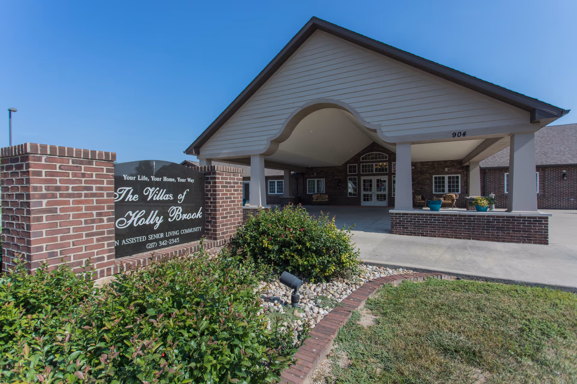 Exterior view of the entrance to The Villas of Holly Brook assisted senior living community, featuring a covered driveway with brick pillars and a sign displaying the facility's name and contact number, surrounded by greenery and landscaping.