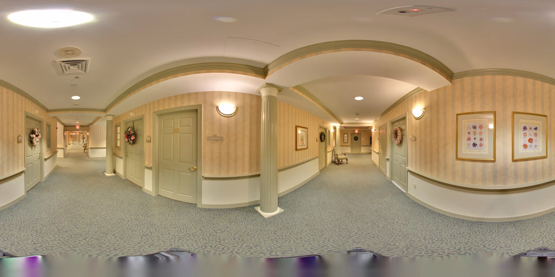 A long, carpeted hallway in an assisted living facility with beige and light green walls, decorated with framed floral artwork and wreaths on the doors. The hallway features columns, wall sconces, and a chair placed against the wall.