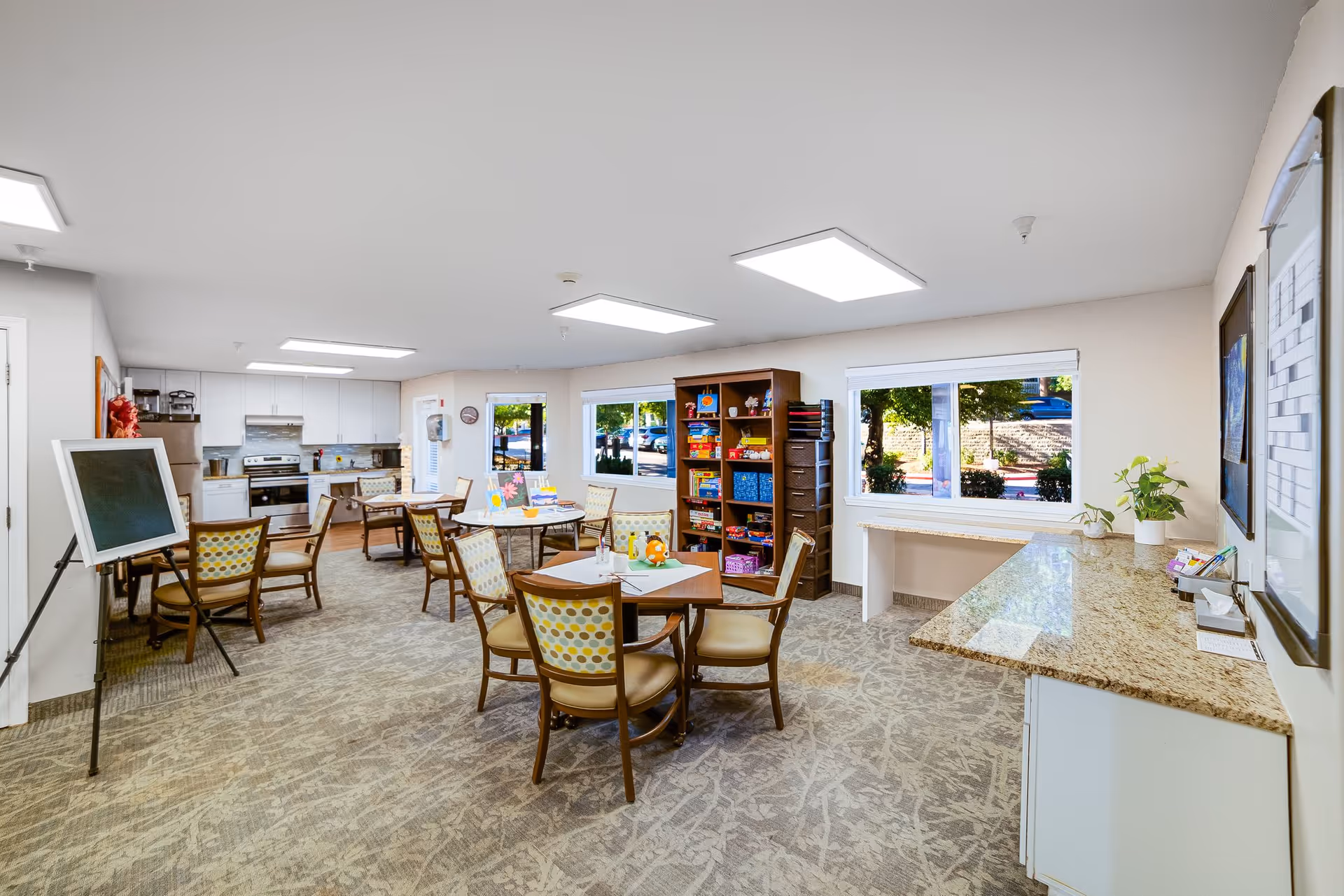 A bright and spacious common room with several tables and chairs arranged for group activities. The room features a kitchen area with white cabinets, a stove, and a refrigerator. There is a bookshelf filled with board games and art supplies, and a chalkboard on an easel. Large windows allow natural light to fill the room, and there are plants and artwork decorating the space.