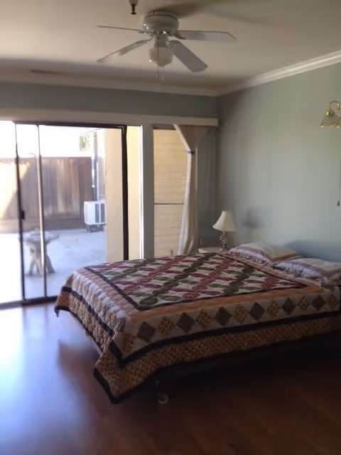 A bedroom with a bed covered in a colorful quilt, two pillows, a small bedside table with a lamp, a ceiling fan above, and a sliding glass door leading to an outdoor patio area.