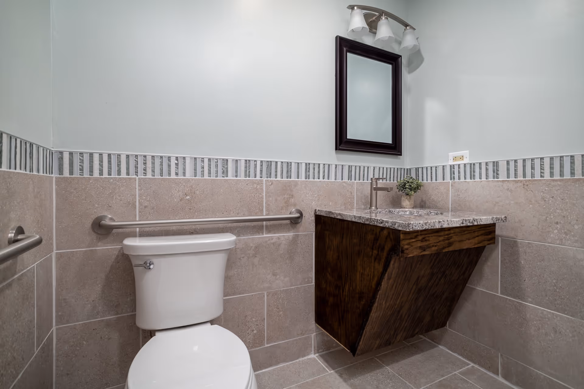 A clean and modern bathroom featuring a white toilet with a horizontal grab bar on the wall behind it, beige tiled walls with a decorative vertical stripe border, a floating wooden vanity with a granite countertop and a small potted plant, a rectangular mirror with a dark frame above the sink, and a three-light fixture mounted above the mirror.