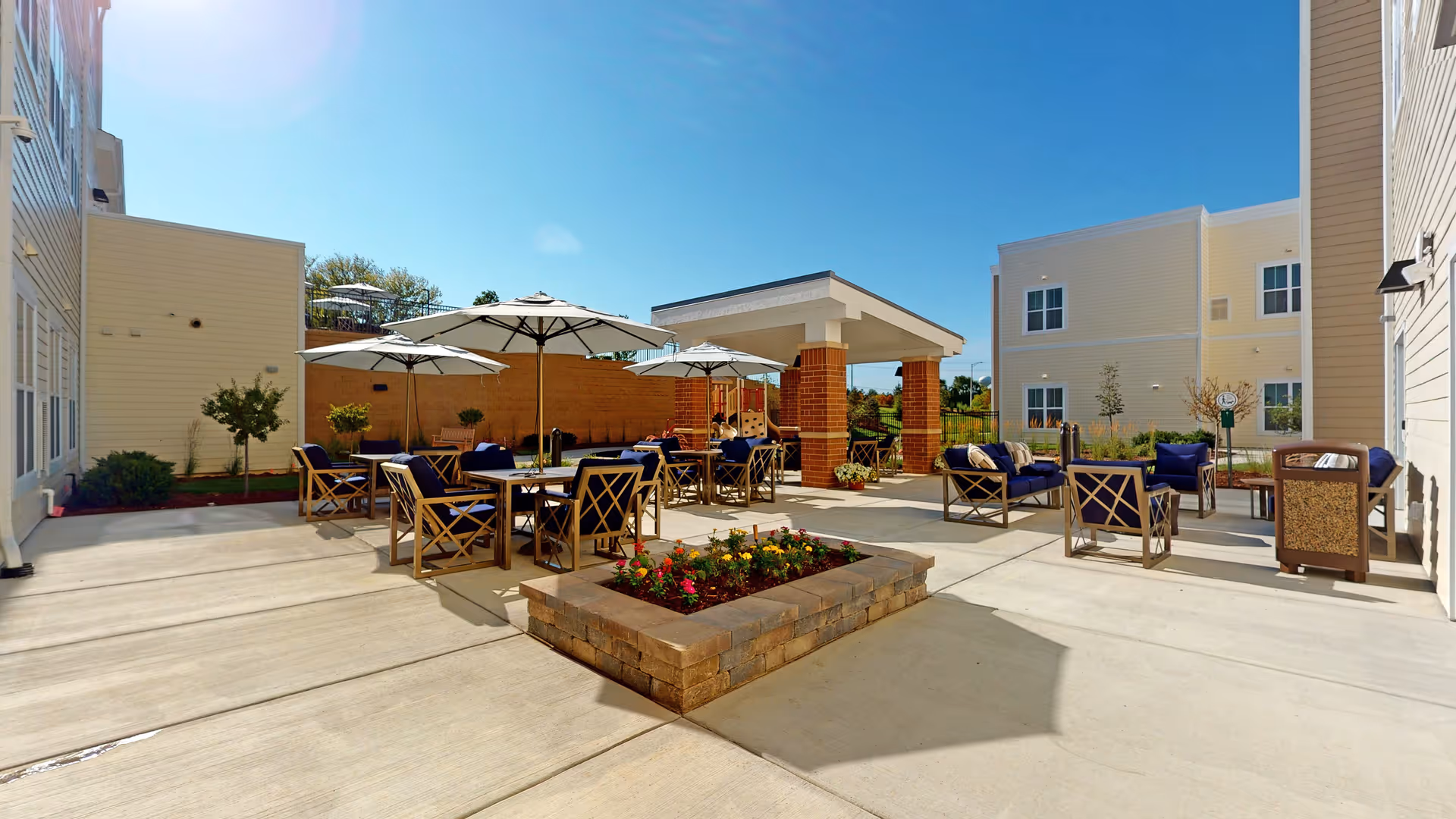 Outdoor patio area at Melody Living Lake in the Hills Assisted Living & Memory Care featuring multiple seating arrangements with tables, chairs, and umbrellas. There is a raised flower bed with colorful flowers in the center, surrounded by concrete flooring. The patio is adjacent to light-colored building walls under a clear blue sky.