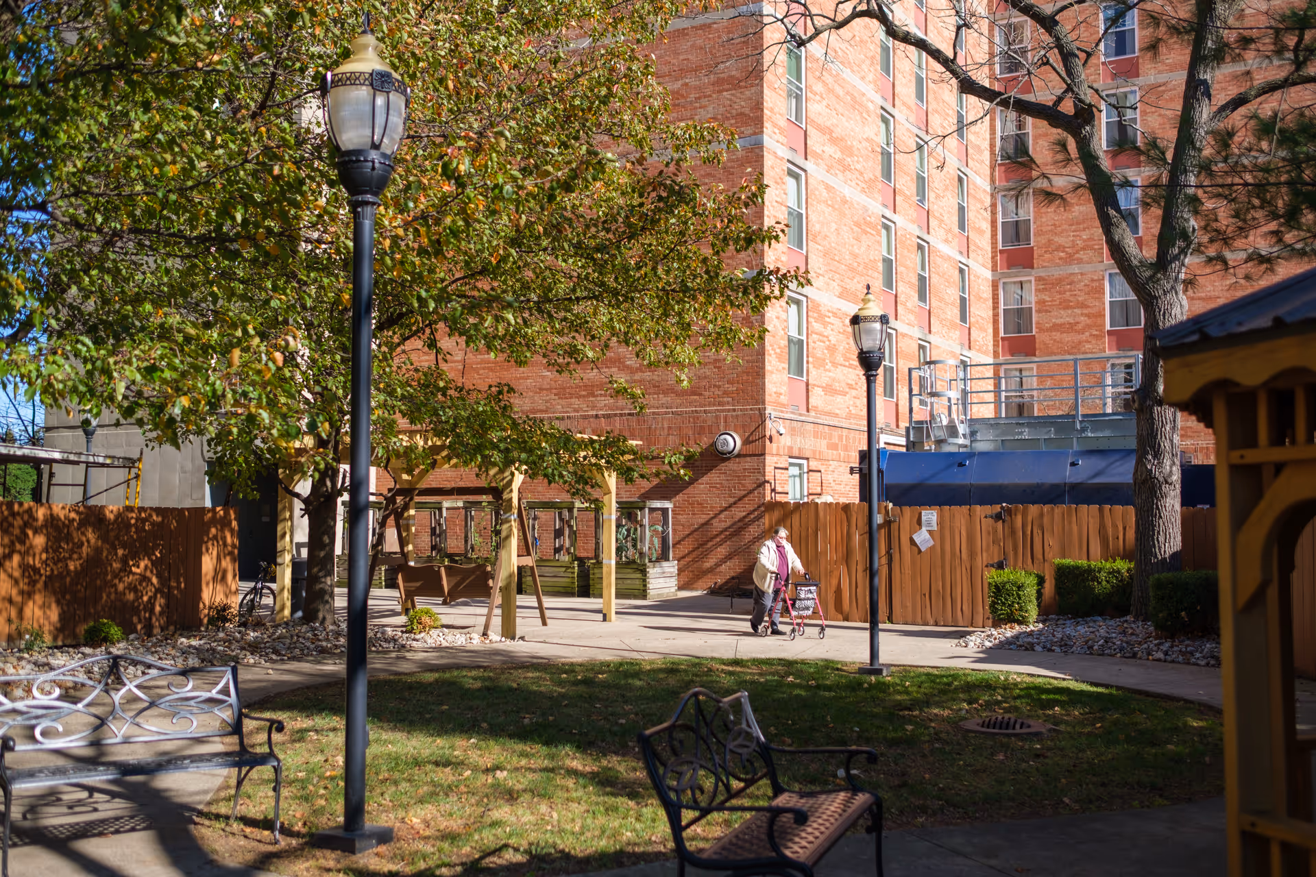 Outdoor courtyard area at Friendship House Louisville with benches, lamp posts, trees, and a person walking with a walker near a brick building.