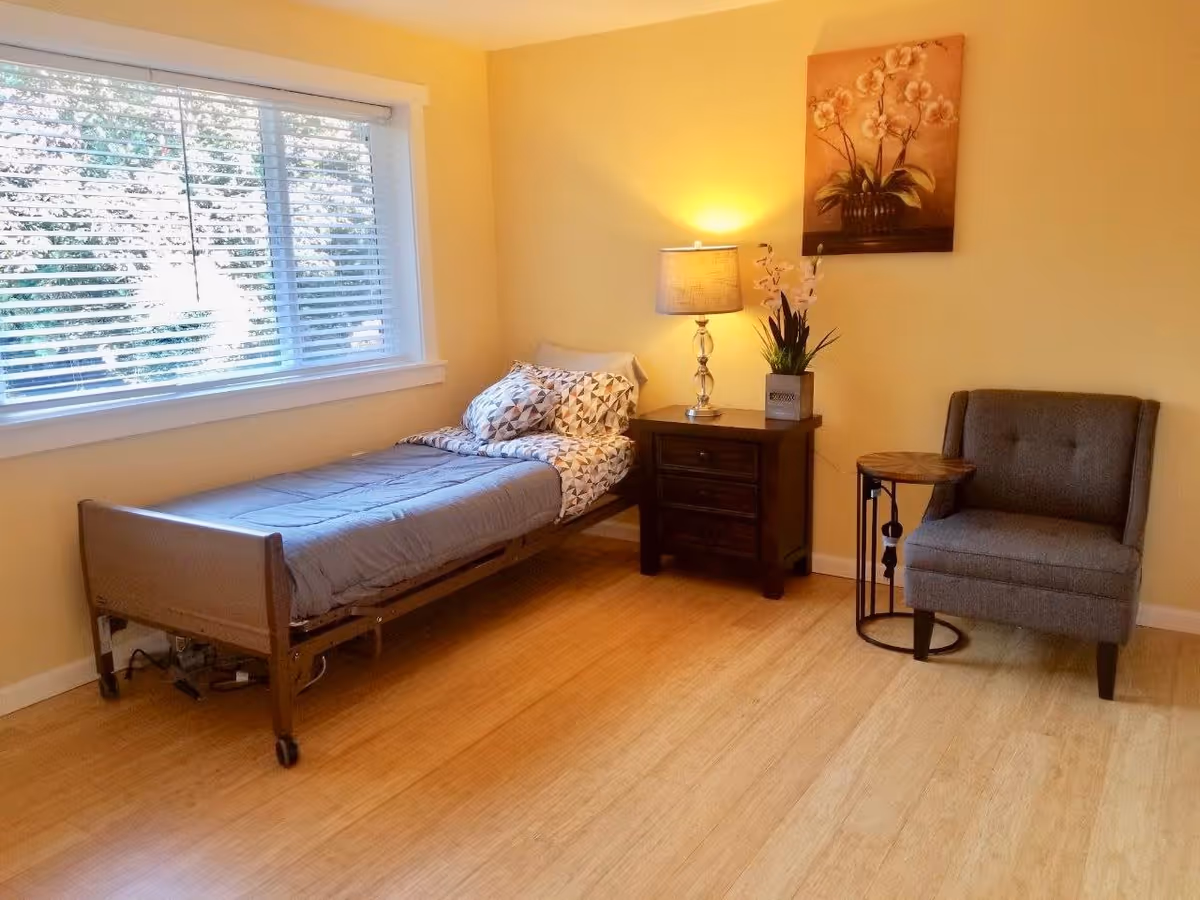 A simple, cozy bedroom with a single bed covered in gray bedding and patterned pillows. Next to the bed is a dark wooden nightstand with a lamp and a potted plant. A gray upholstered chair and a small round side table are placed against the wall. A painting of flowers hangs above the nightstand. The room has light-colored walls and a large window with blinds, letting in natural light. The floor is wooden.