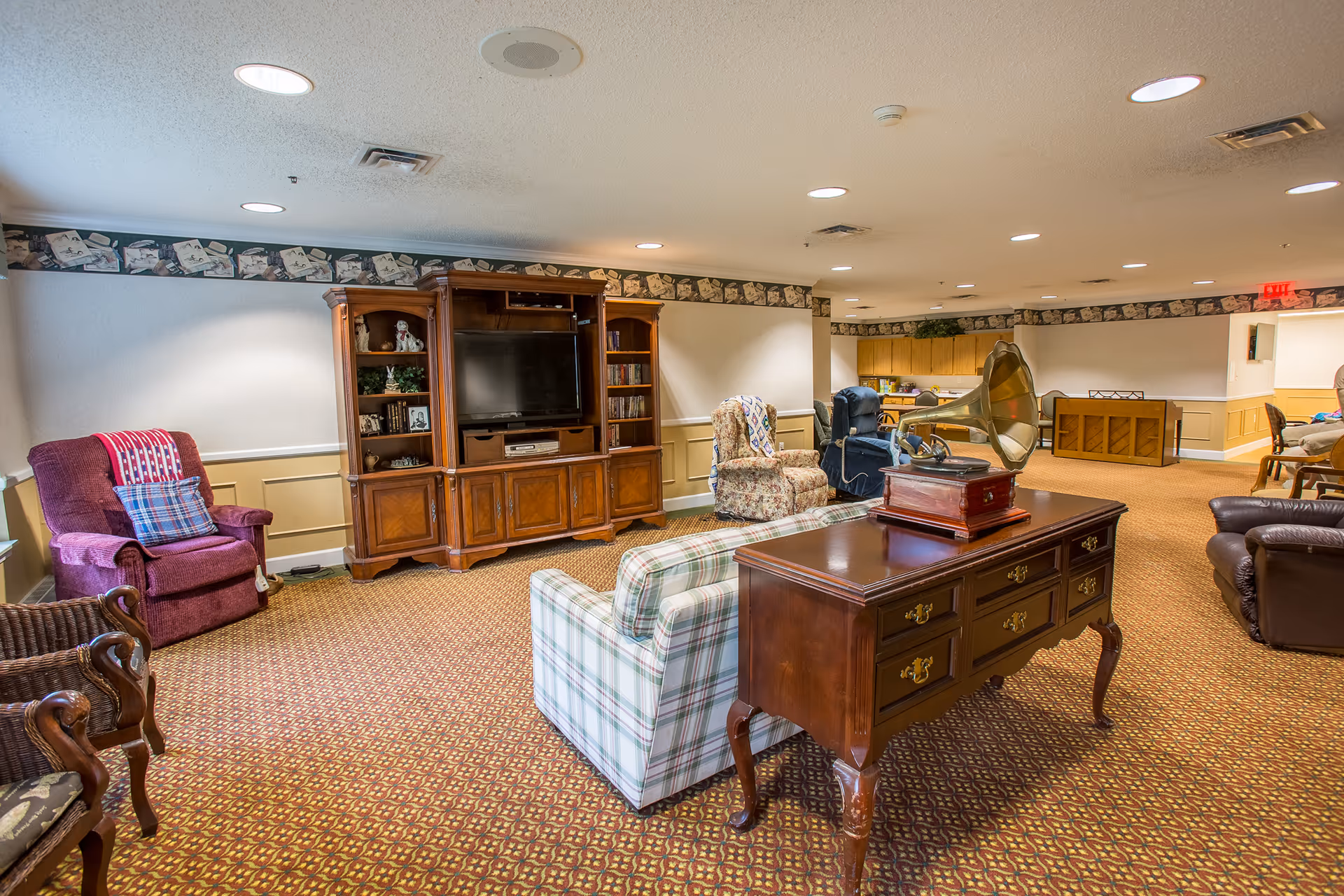 A cozy senior living facility common area with various upholstered chairs, a wooden entertainment center with a TV, a vintage gramophone on a wooden table, and a piano in the background. The room has patterned carpet, wallpaper border near the ceiling, and recessed lighting.