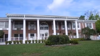 Two-story brick building with large white columns, a central entrance and a manicured lawn under a blue sky.