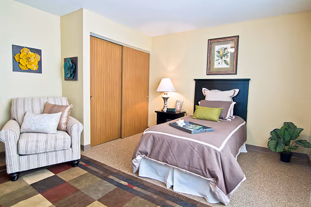 Well-lit bedroom with a single bed, nightstand and lamp, an armchair, sliding closet doors, and decorative wall art.