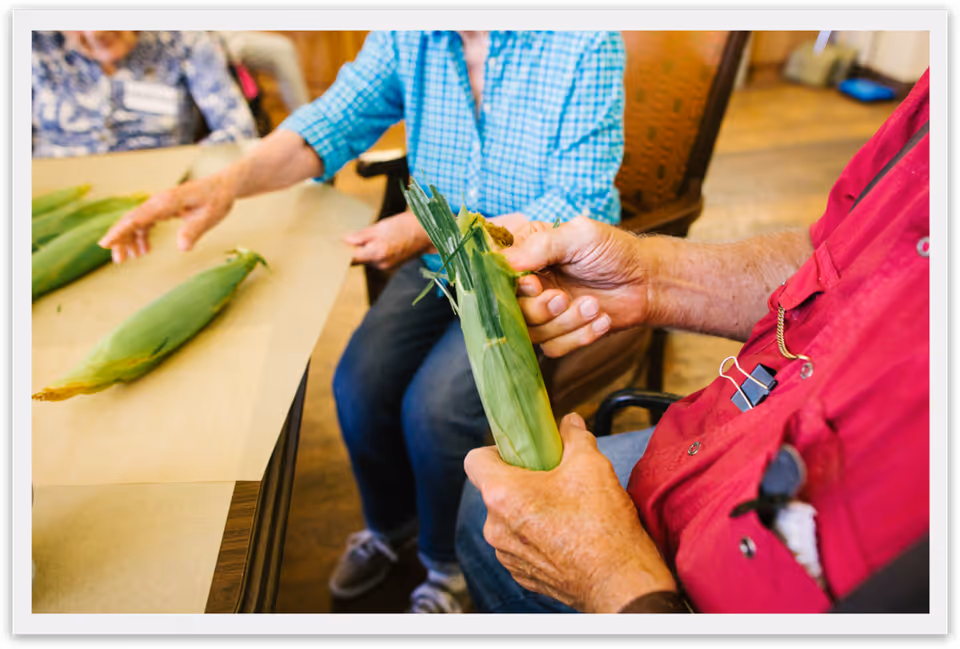 Two elderly individuals sitting at a table, one wearing a red shirt and the other a blue checkered shirt, husking ears of corn. The setting appears to be a communal indoor space with wooden flooring and chairs.
