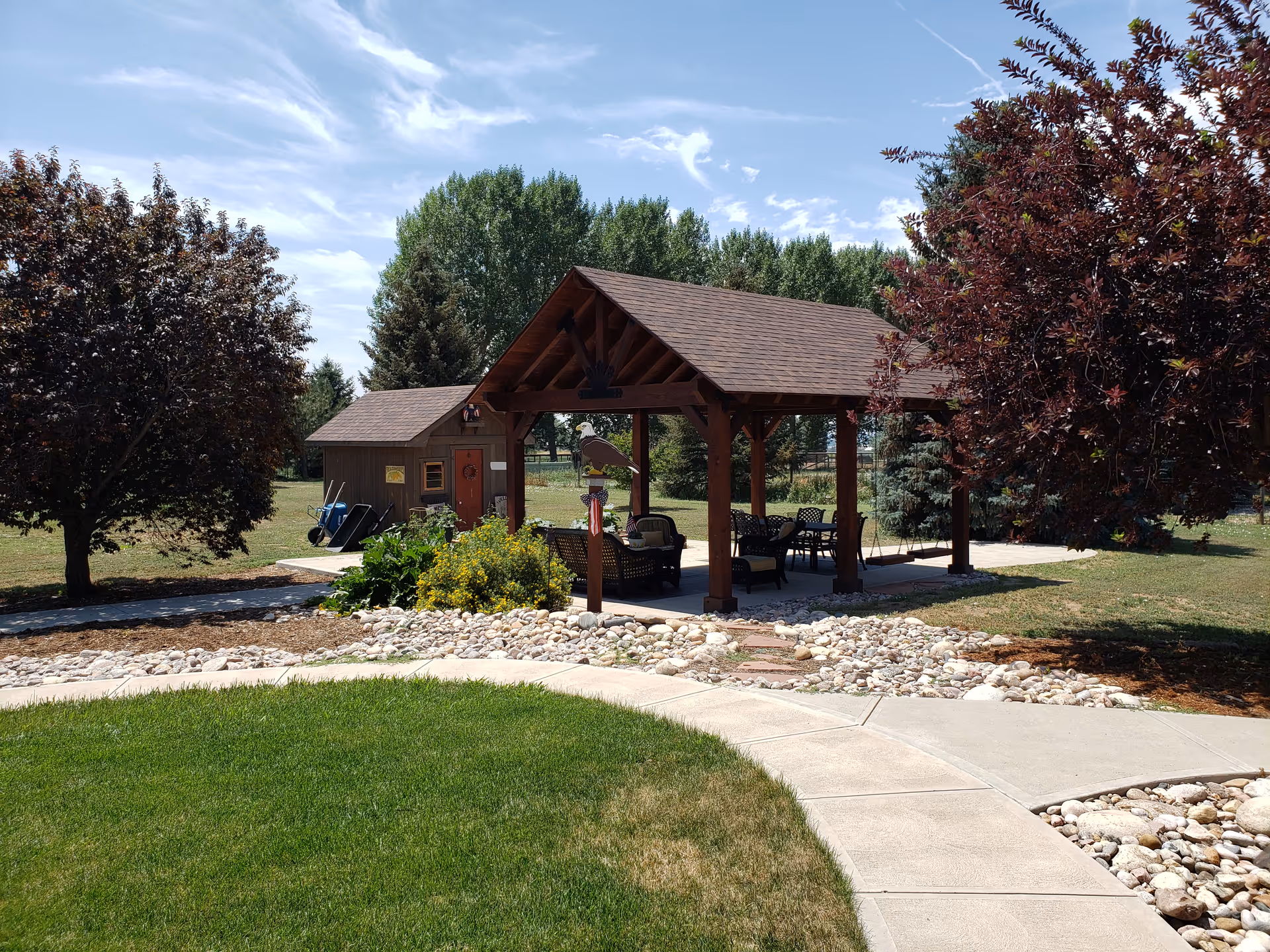 Wooden pavilion with seating and a small shed in a landscaped lawn with trees and walkways.
