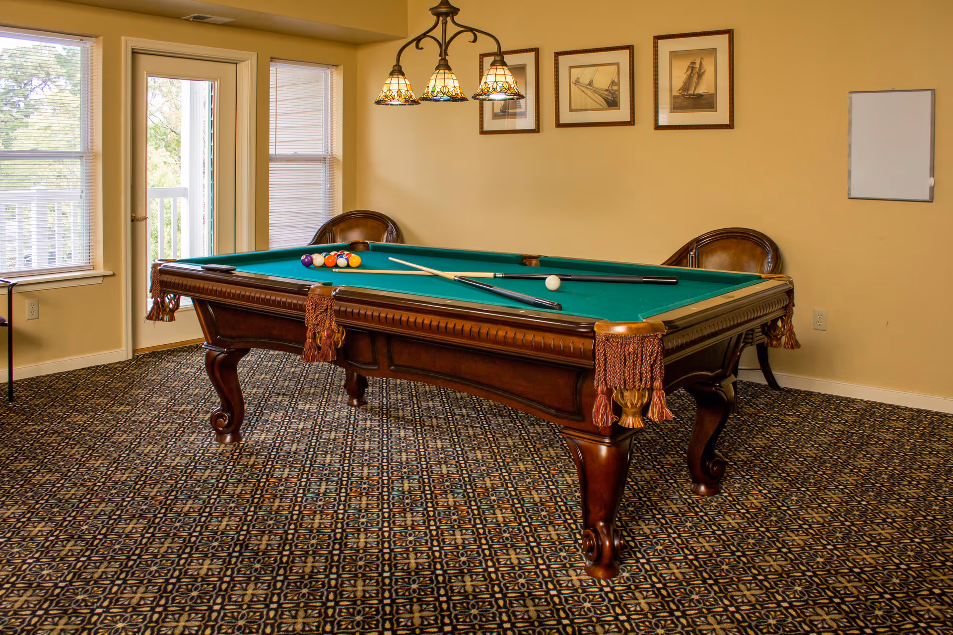 A room with a classic wooden pool table with green felt, pool cues, and balls arranged on it. The room has patterned carpet flooring, beige walls, three framed pictures of sailboats on the wall, two wooden chairs, a hanging light fixture with three lamps above the pool table, and a door and windows letting in natural light.