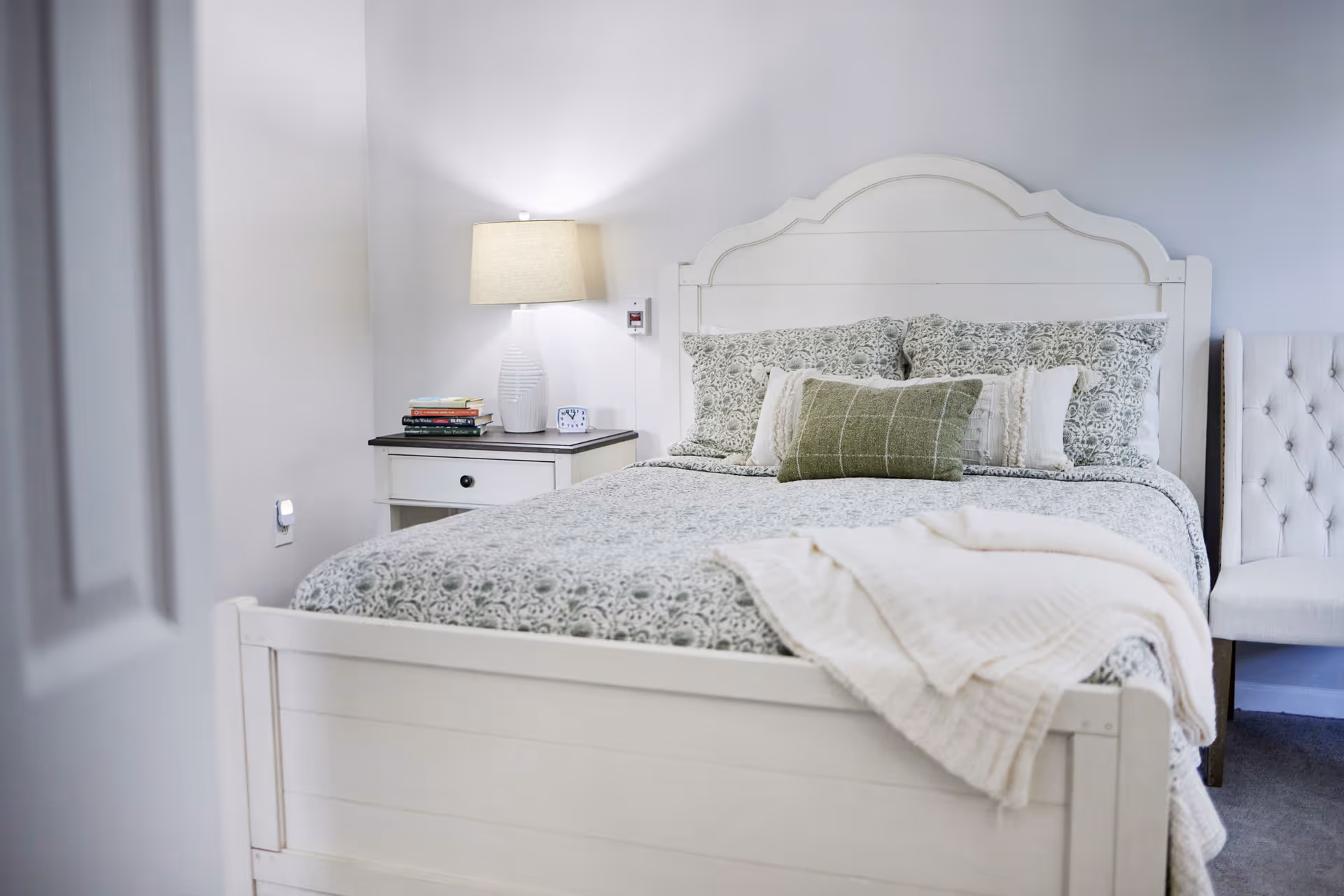 Bright bedroom with a white wooden bed dressed in patterned bedding, a nightstand with a lamp and books, and an upholstered chair.