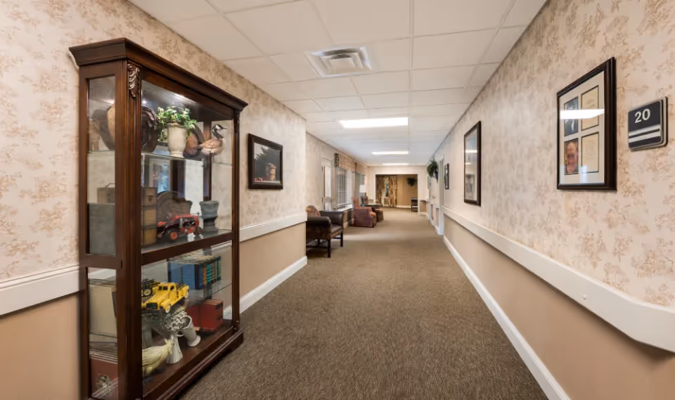 A long, carpeted hallway in a senior living facility with beige floral wallpaper and white trim. On the left side, there is a wooden display cabinet with glass doors containing decorative items and books. Several framed pictures hang on the walls, and there are chairs placed along the hallway. The ceiling has recessed lighting panels.